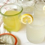 Two glasses of lemonade made with Classic Lemonade Syrup are garnished with lemon slices, sitting on a table beside a jar of lemonade, its lid, and a spoon on a white towel.