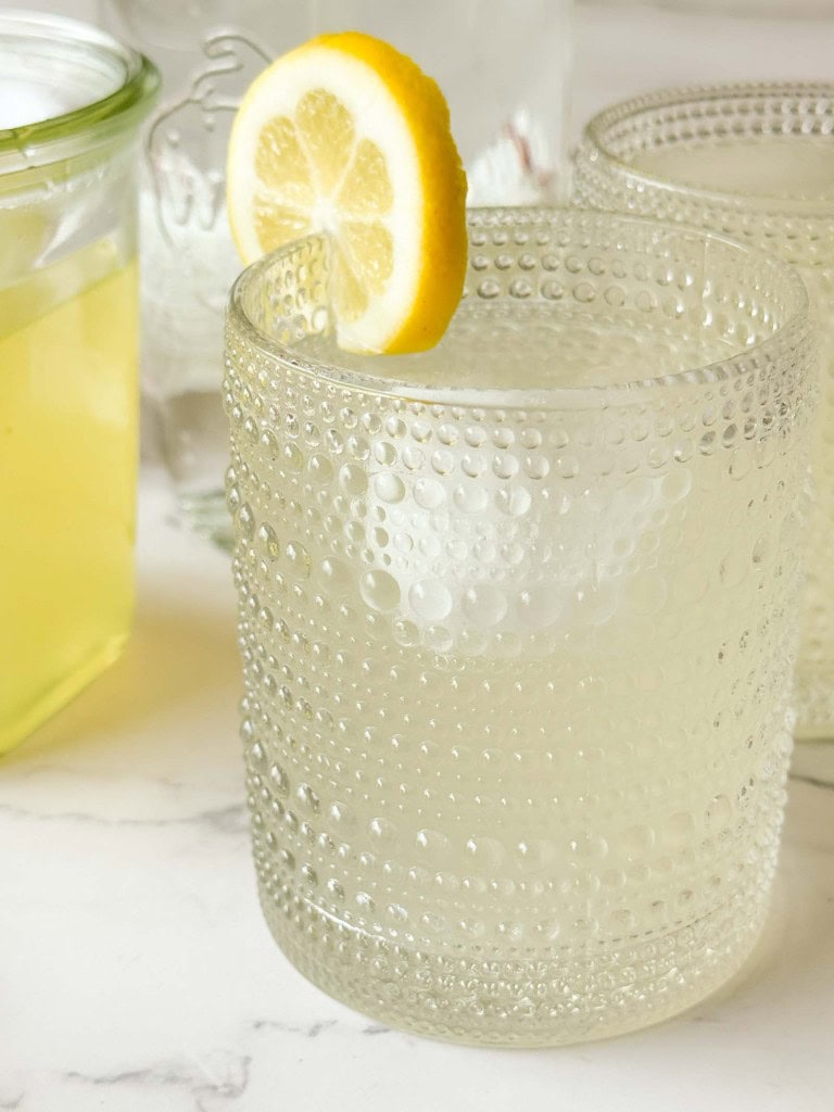 A textured glass filled with a clear lemon drink and ice, made using Classic Lemonade Syrup and garnished with a slice of lemon on the rim, sits on a white surface.