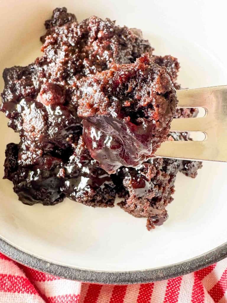 A close-up of a fork holding a portion of chocolate cobbler with a gooey, glossy chocolate sauce on a white plate.