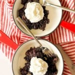 Two white bowls with chocolate dessert topped with whipped cream, each with a spoon, placed on a red and white striped cloth.