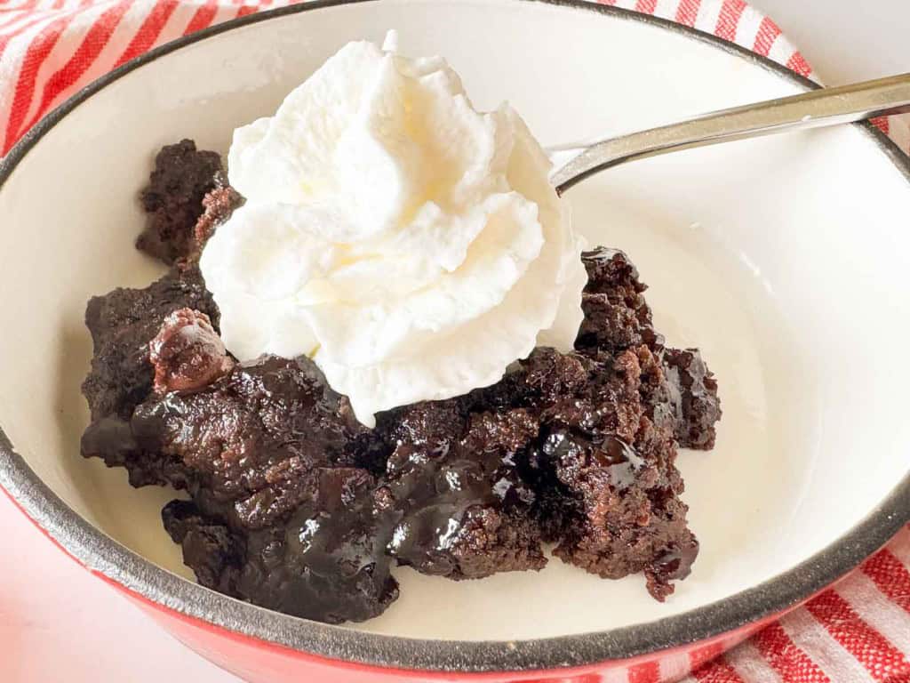 A bowl of chocolate lava cake topped with whipped cream, served on a white plate with a spoon and a red-striped napkin in the background.