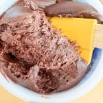 A yellow spatula scooping thick chocolate frosting from a white container on a light wooden surface.