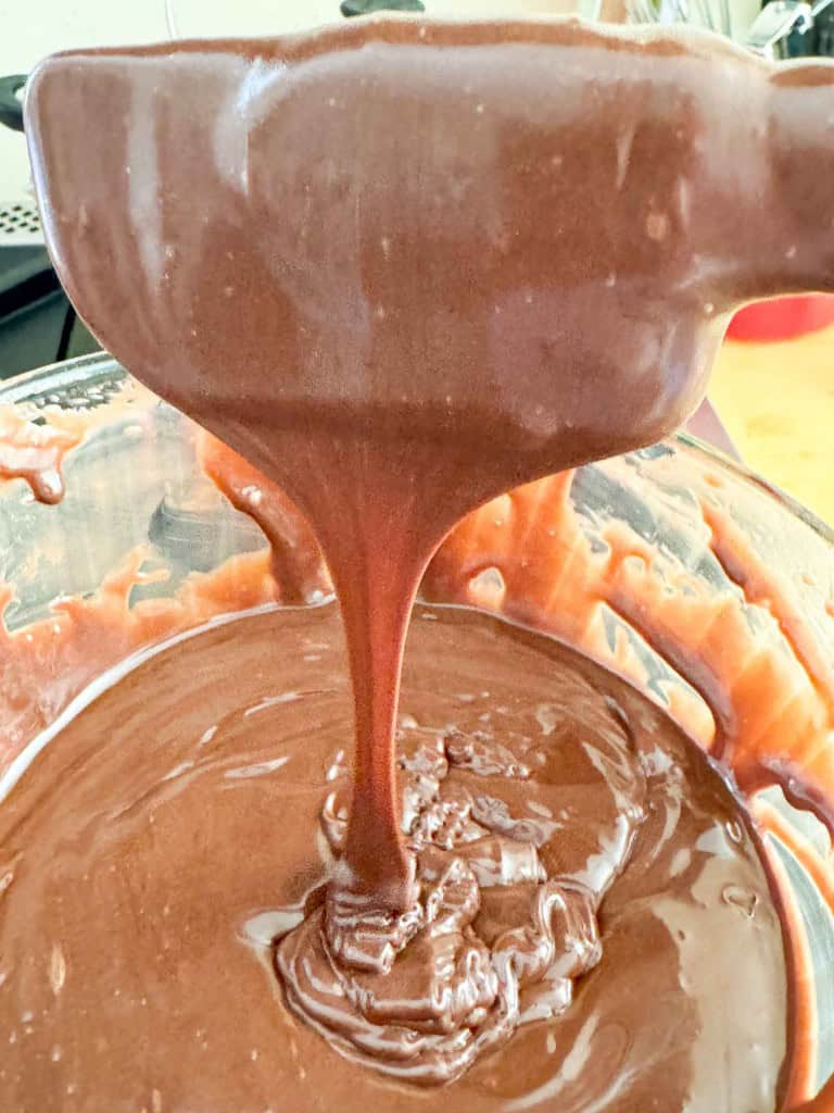 A spoon drizzling thick chocolate batter into a glass mixing bowl filled with more batter.