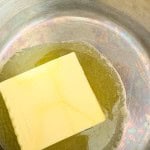A rectangular piece of butter is partially melted in a stainless steel pan.