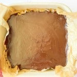 A square baking dish lined with parchment paper, filled with a smooth layer of chocolate over a dessert, placed on a red and white checkered towel.