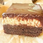 A close-up of a layered dessert bar with a chocolate base, creamy middle layer, and glossy chocolate topping, placed on parchment paper.