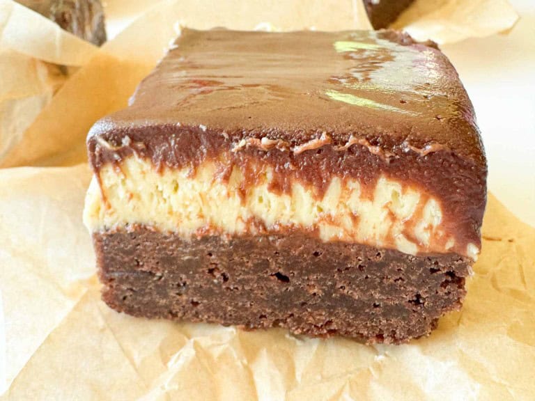 A close-up of a layered dessert bar with a chocolate base, creamy middle layer, and glossy chocolate topping, placed on parchment paper.
