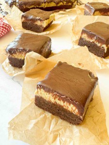 Squares of chocolate brownies with a cheesecake layer and chocolate topping are placed on pieces of brown parchment paper.