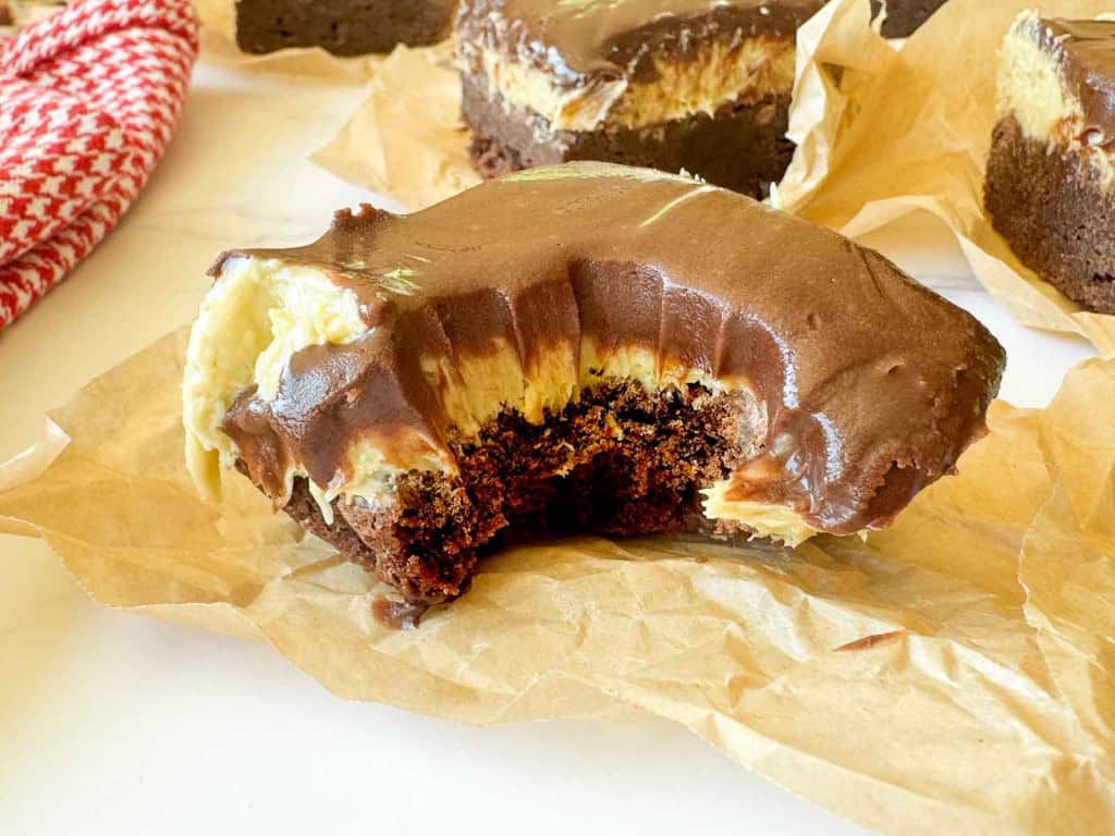 A chocolate and cream layered brownie bar with a large bite taken out, resting on a piece of parchment paper.