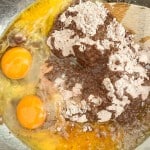 A mixing bowl contains two raw eggs, brownie mix powder, oil, and water, with a wooden spoon partially visible.