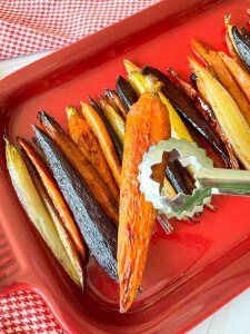 Tongs holding a roasted carrot on a red tray with rows of roasted multicolored carrots arranged neatly.