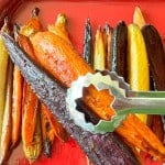 A metal tong grips a roasted purple carrot among other assorted colorful roasted carrots on a red tray.