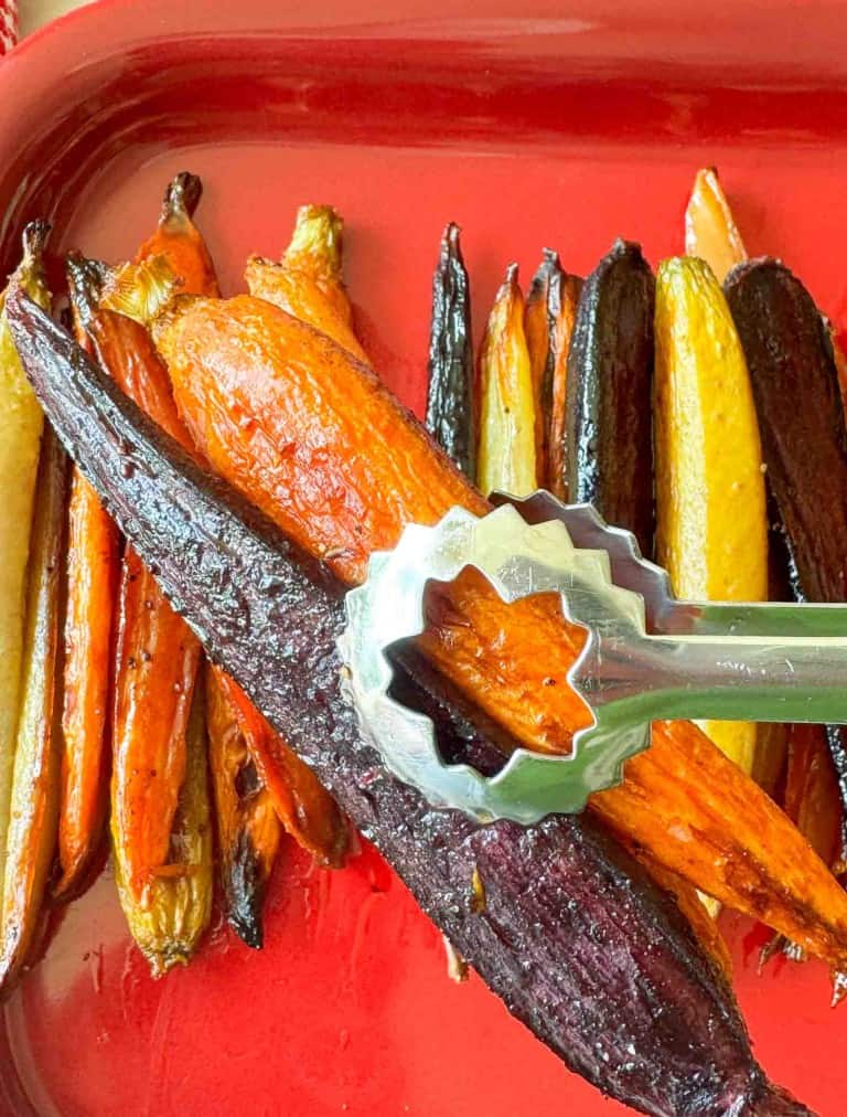 A metal tong grips a roasted purple carrot among other assorted colorful roasted carrots on a red tray.