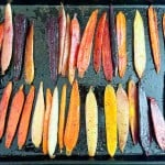 Colorful carrot slices, including orange, purple, and yellow varieties, arranged in rows on a black baking tray ready for roasting.
