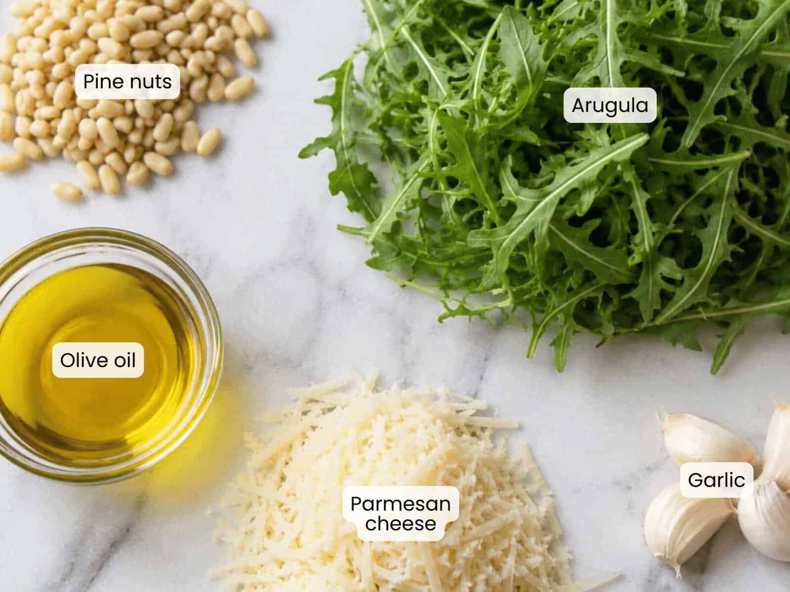 A flat lay of pine nuts, arugula, olive oil, grated Parmesan cheese, and garlic&mdash;each labeled and neatly arranged on a white surface&mdash;showcases the fresh ingredients for classic arugula pesto.