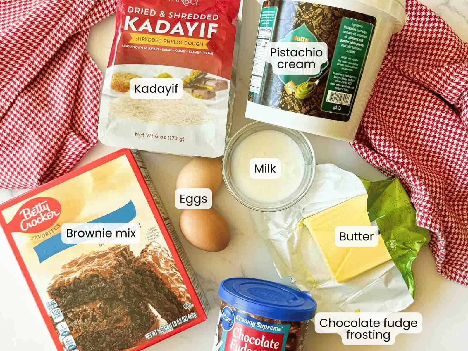 A flat lay of ingredients including kadayıf, pistachio cream, eggs, milk, butter, brownie mix for Dubai chocolate brownies, and chocolate fudge frosting on a red checkered cloth.