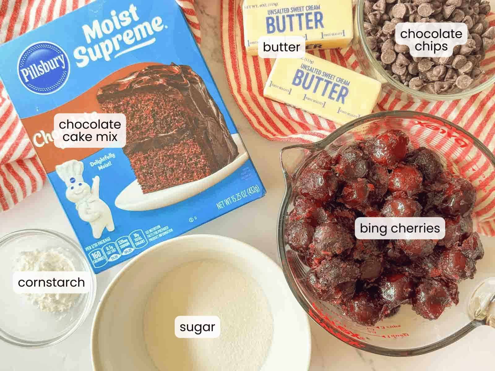 Ingredients for a Slow Cooker Cherry ChocolateCobbler are displayed: box of chocolate cake mix, bowl of bing cherries, chocolate chips, two sticks of butter, sugar, and cornstarch&mdash;each one clearly labeled on the countertop.