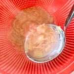 A metal ladle holds some shredded cooked vegetables, perhaps for a refreshing rhubarb lemonade, over a red plastic strainer, with liquid draining into a white container below.