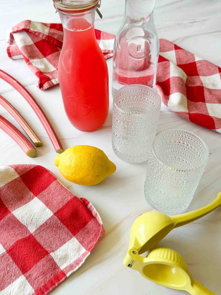 A bottle of pink rhubarb lemonade, a clear carafe, two empty glasses, a lemon, rhubarb stalks, a yellow lemon squeezer, and red plaid towels on a white surface.