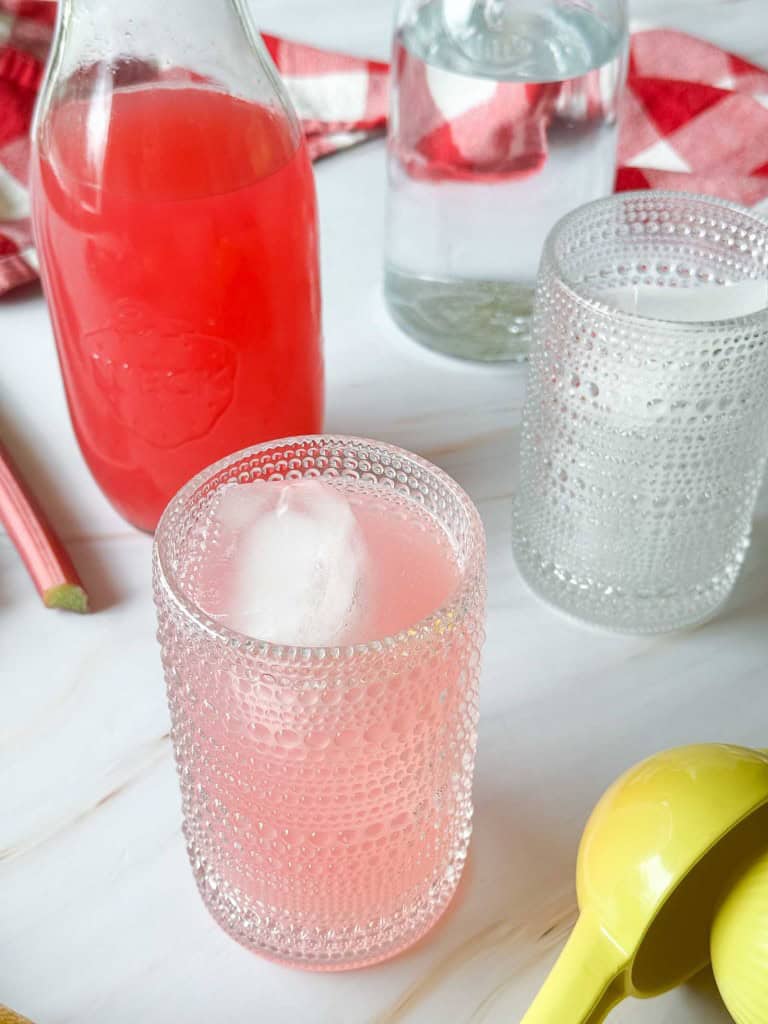 A textured glass filled with pink rhubarb lemonade and ice sits on a white surface, next to a clear glass, a glass bottle of pink liquid, and a yellow citrus juicer.