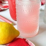 A textured glass filled with rhubarb lemonade sits on a red checkered cloth next to a lemon, with a bottle of pink drink in the background.