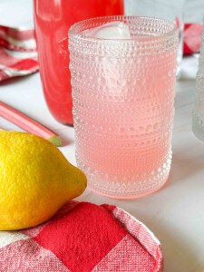 A textured glass filled with rhubarb lemonade sits on a red checkered cloth next to a lemon, with a bottle of pink drink in the background.