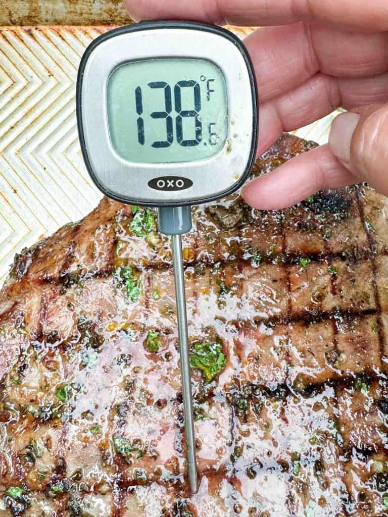 A hand holds a digital meat thermometer reading 138&deg;F, inserted into a grilled steak on a tray.