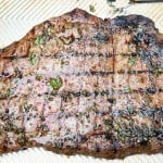Grilled steak with visible char marks, seasoned with herbs and juices, resting on a light-colored grooved surface.