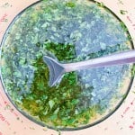 A metal spoon stirs a green herb and liquid mixture in a glass measuring cup, viewed from above.