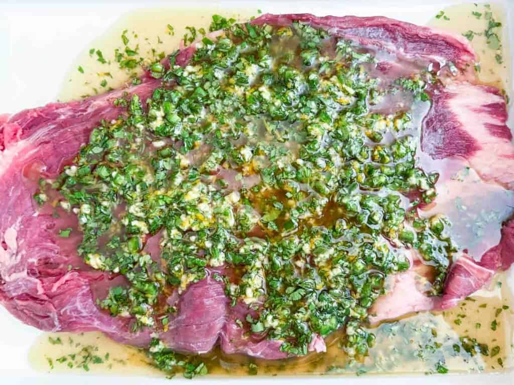 Raw flank steak topped with a marinade of chopped herbs, garlic, and oil, displayed on a white surface.
