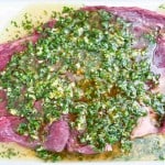 Raw flank steak topped with a marinade of chopped herbs, garlic, and oil, displayed on a white surface.