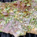 Raw marinated flank steak with herbs and seasoning placed on a grill, ready to be cooked.