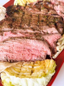Close-up of grilled steak, sliced to show a pink, medium-rare center, served on a red plate with charred wedges of cabbage.