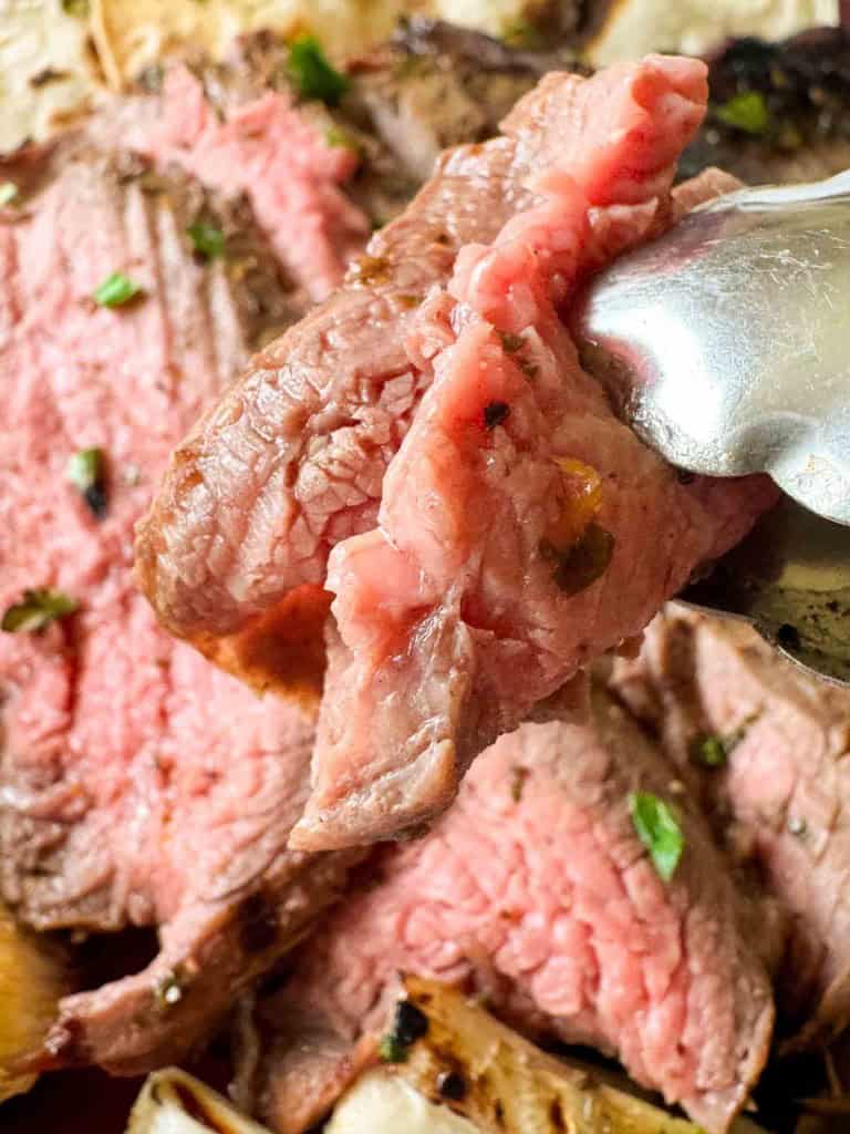 Close-up of a metal fork holding a slice of medium-rare beef steak, with more sliced steak in the background.