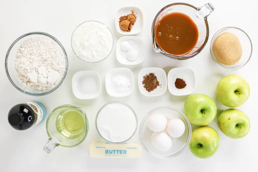 Top-down view of baking ingredients for an apple spice cake, including flour, eggs, apples, brown sugar, oil, vanilla, yogurt, butter, white sugar, caramel sauce, baking powder, spices, baking soda, and salt.