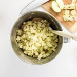 A mixing bowl filled with diced apples and batter for an apple spice cake, a wooden spoon, a cutting board with sliced apples, a whole green apple, and a measuring cup with brown liquid on a white surface.