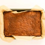 A rectangular loaf of baked banana bread, reminiscent of an apple spice cake with its warm, inviting aroma, sits in a parchment paper-lined metal pan on a white background.