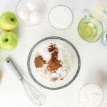 Top-down view of baking ingredients for an apple spice cake, including green apples, eggs, sugar, flour with spices, oil, brown sugar, butter, vanilla, sour cream, and caramel sauce arranged on a white surface.