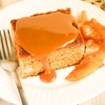 A slice of apple spice cake topped with caramel sauce is served on a white plate with cooked apple slices and a fork.