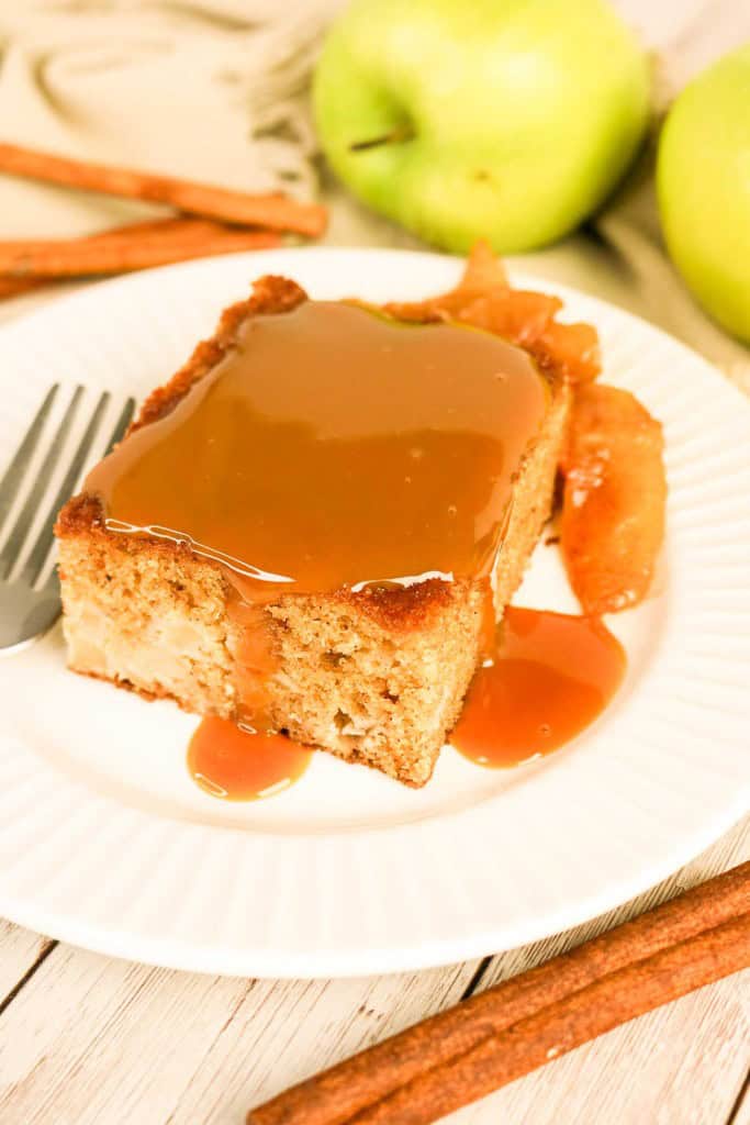 A slice of apple spice cake with caramel sauce and baked apple slices sits on a white plate, next to a fork, two green apples, and cinnamon sticks.