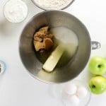 Mixing bowl with butter, brown and white sugar, surrounded by eggs, green apples, flour, sour cream, caramel sauce, and vanilla on a white surface—everything you need to create a delicious apple spice cake.