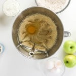 Top-down view of baking ingredients for apple spice cake, including an egg in a mixing bowl, apples, eggs, flour, yogurt, caramel sauce, and vanilla extract on a white surface.