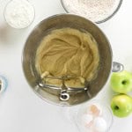 Stand mixer bowl with batter for apple spice cake, surrounded by green apples, eggshells, dry ingredients, vanilla extract, caramel sauce, and yogurt on a white surface.
