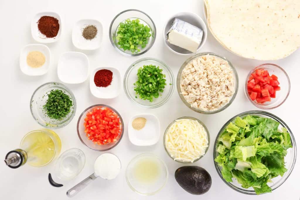Various chopped vegetables, seasonings, shredded cheese, cooked chicken, and tortillas are arranged in glass bowls and plates on a white surface&mdash;perfect for assembling delicious Firecracker Chicken Wraps.