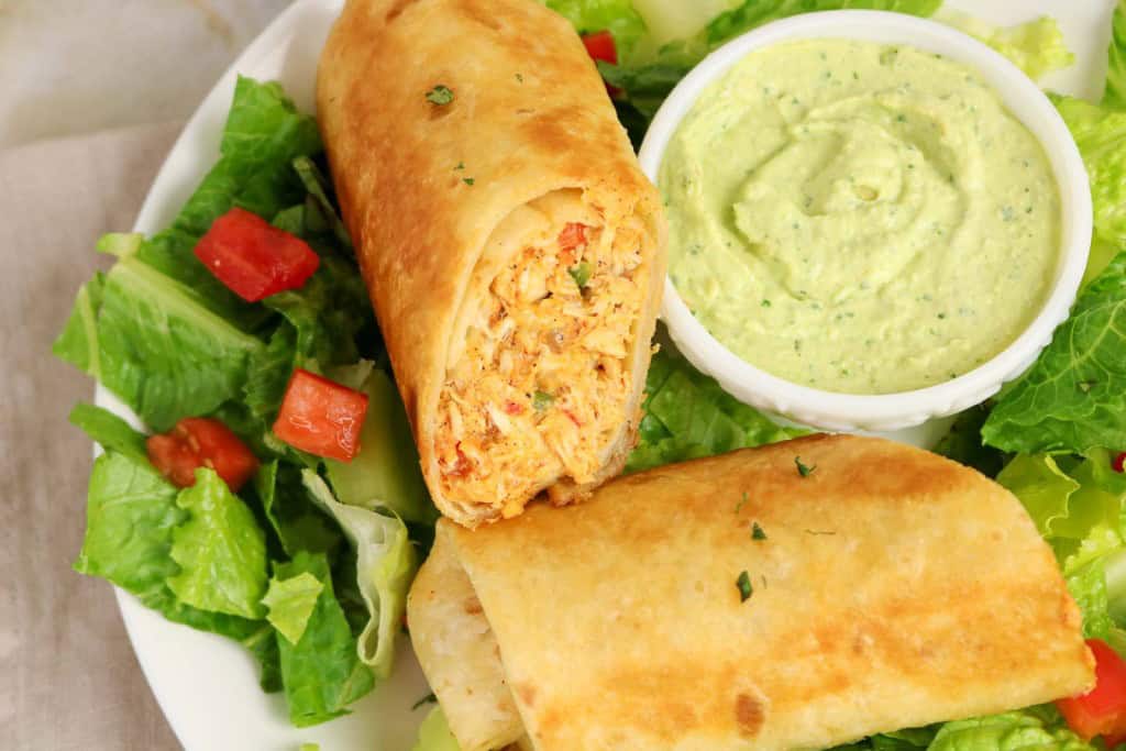 A plate with a cut chicken chimichanga, reminiscent of Firecracker Chicken Wraps, served alongside a green salad with diced tomatoes and a small cup of creamy green sauce.