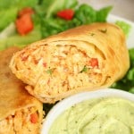 A close-up of two halved, crispy chicken chimichangas&mdash;reminiscent of Firecracker Chicken Wraps&mdash;served on lettuce with diced tomatoes, next to a bowl of creamy green avocado dip.
