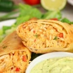 A close-up of a halved Firecracker Chicken Wraps burrito with shredded filling, served on lettuce with a side of creamy green dip.