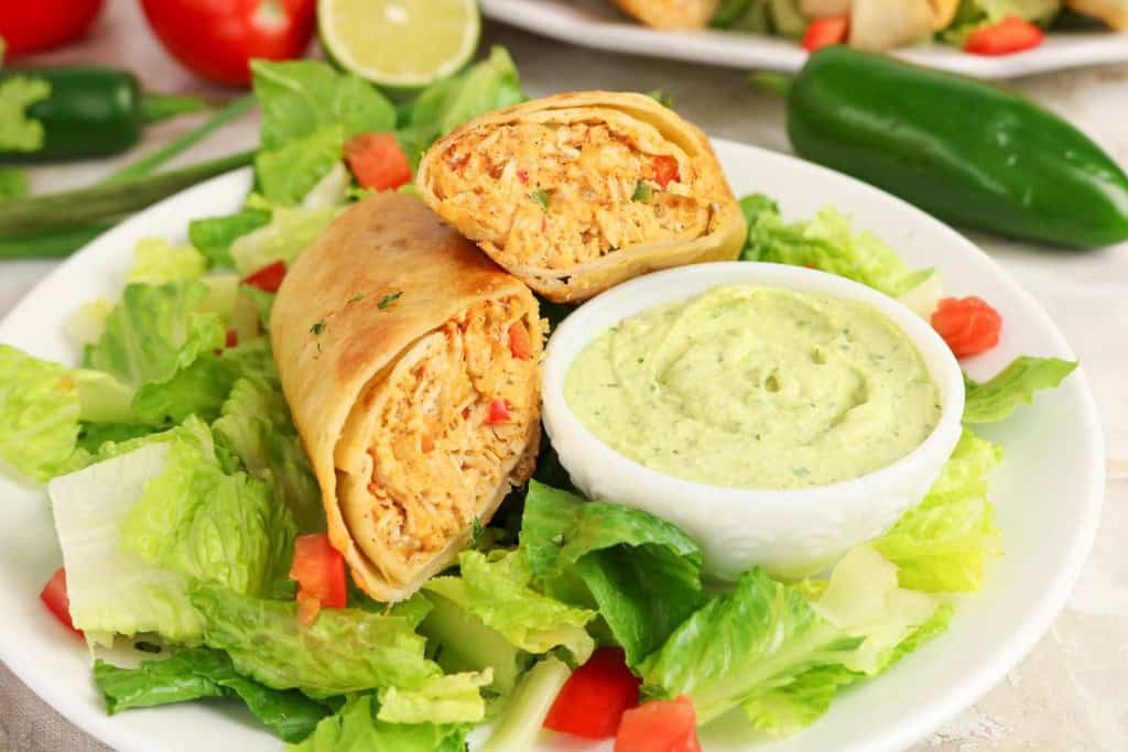 A burrito cut in half served on lettuce with diced tomatoes, reminiscent of flavorful Firecracker Chicken Wraps, sits next to a small bowl of creamy green sauce on a white plate.