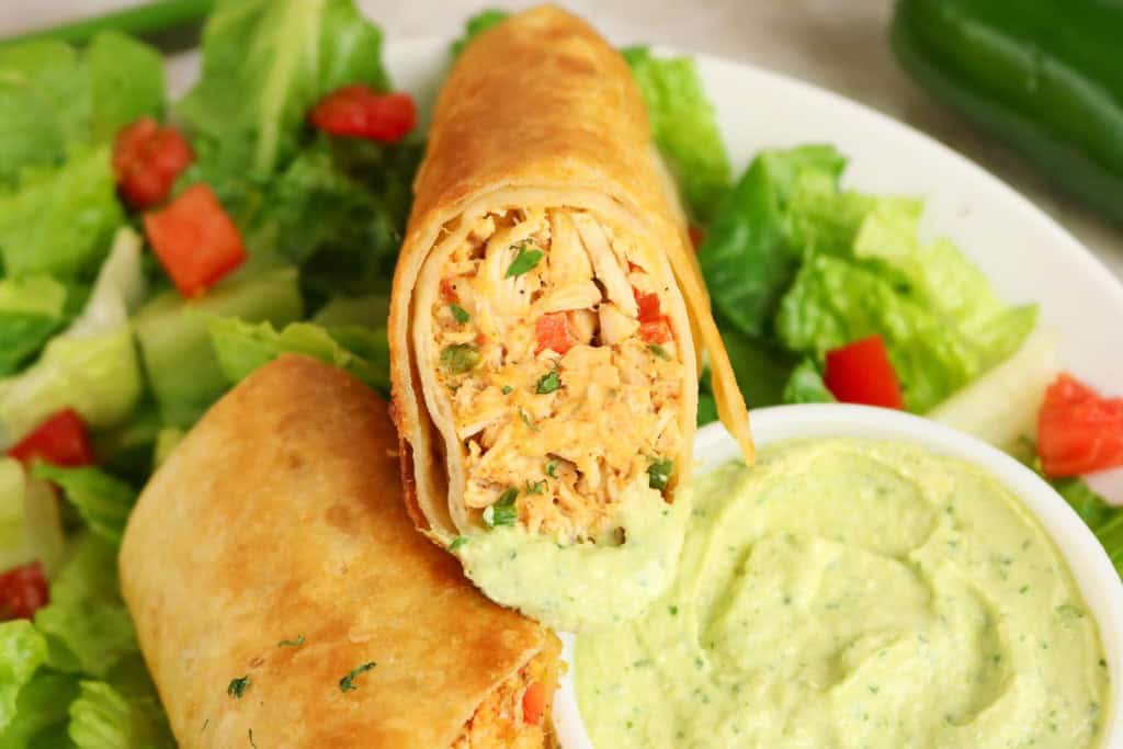 A close-up of a chicken chimichanga cut in half to reveal the shredded filling, reminiscent of Firecracker Chicken Wraps, served with a side of green dipping sauce and a fresh green salad with diced tomatoes.