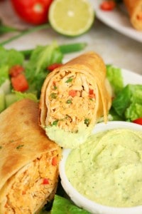 A close-up of a halved, crispy chicken chimichanga with a creamy green dipping sauce, served on a bed of lettuce with tomatoes and a lime in the background, reminiscent of the bold flavors found in Firecracker Chicken Wraps.